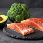 Полезные продукты для уменьшения живота: авокадо, брокколи и лосось на каменной столешнице.